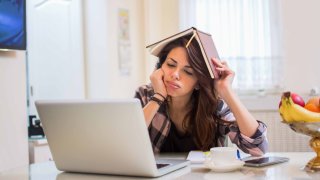 Foto ilustrativa de una estudiante en una computadora portátil y sosteniendo un cuaderno en la cabeza.