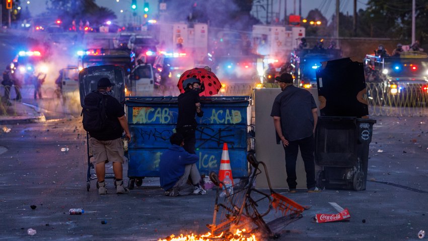 COMPTON, CA -JUNE 7, 2025: Protesters hide behind a dumpster near a set  fire as  LA County Sheriff deputies shoot projectiles to keep demonstrators from advancing  after ICE raids at a nearby Home Depot and the Garment District brought out resistance from Los Angeles residents on June 7, 2025 in Compton, California.(Gina Ferazzi / Los Angeles Times via Getty Images)